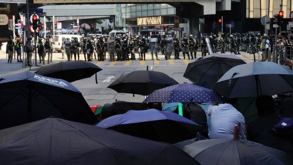 Purtret da demonstrants che tegnan parasols entamaun, visavi ad els stattan duas colonnas da polizists e quai amez la citad da Hongkong. 