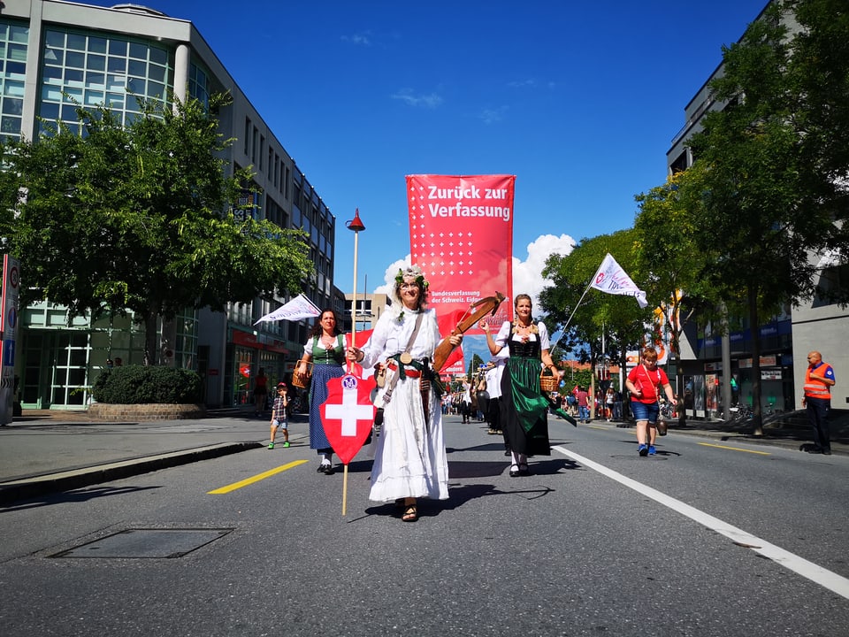 Ina dunna costumada sco Helvetia, davos ella vegn purtada ina bandiera cun scrit: «Zurück zur Verfassung»