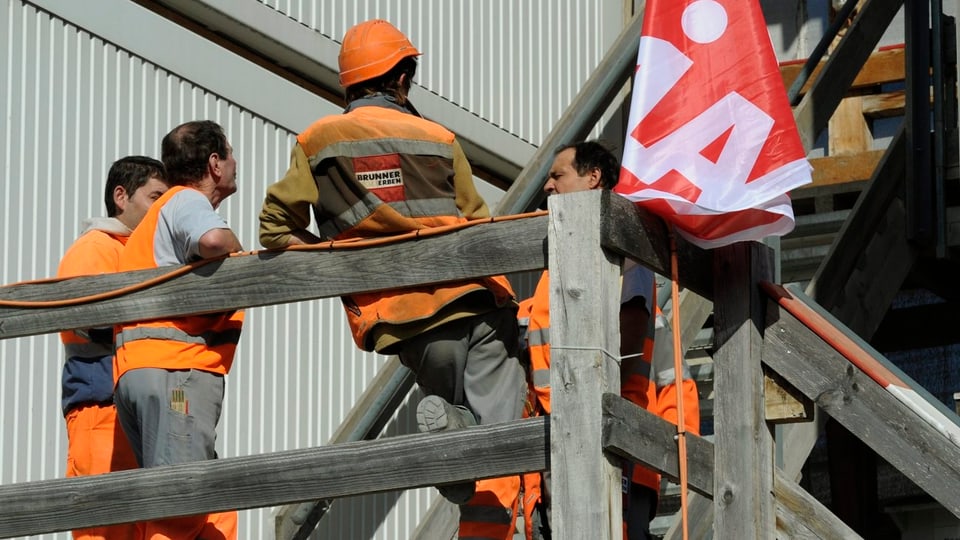 Purtret da lavurers che stattan enturn sin in plazzal. Ina bandiera dal sindicat UNIA penda dasperas. 