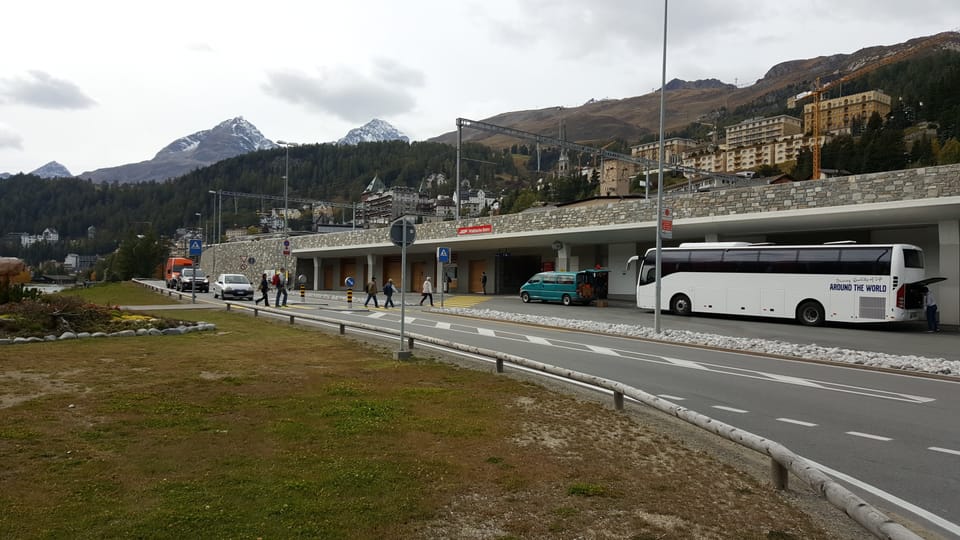 Sguard sin la strivla melna tranter la staziun da San Murezzan e la riva dal lai. Sin la strivla melna hai glieud, davant spetgan autos ed a dretga sin il parcadi da bus è in bus alv.
