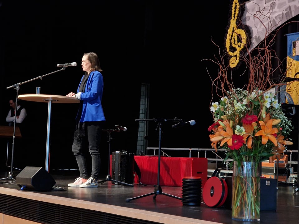 Nora Saratz Cazin trägt schwarze Hosen und einen blauen Blazer und steht auf einer Bühne an einem Rednerpult.