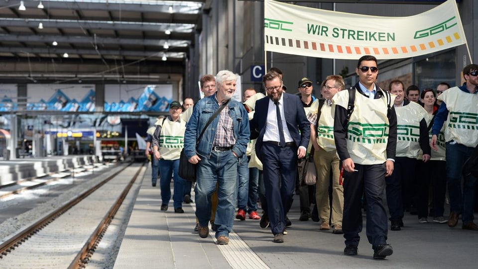 Umens chaminan tras ina staziun da tren cun ina bandiera Wir Streiken