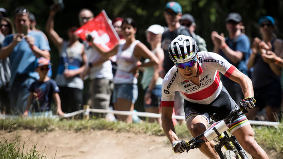 Nino Schurter sin in mountain bike en ina curva. Il terren e natiral e schetg. davostiers ina gruppa d'aspectaturs che applaudeschan. Ina bandiera svizra è da vesair.