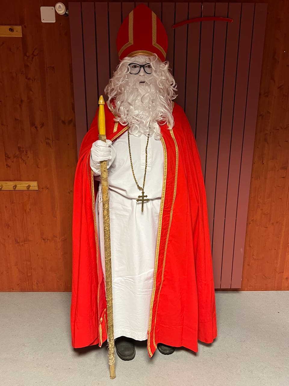 Weihnachtsmann in seiner vollen Montur posiert für ein Bild. Rote Robe, weisses Kleid, Stock, weisse Haare und Bart, Hut