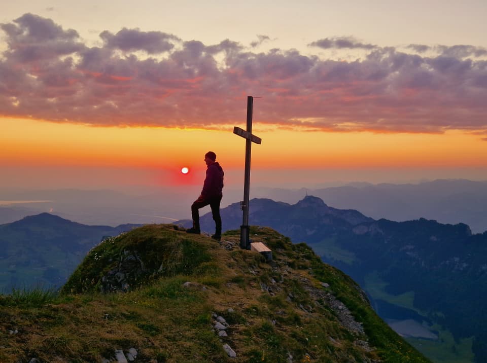 Mann auf Berg neben Kreuz bei Sonnenuntergangsstimmung