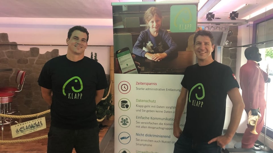 Reto Kaspar ed Elias Schibli Dus umens cun t-shirts nairs cul medem logo, sper in plakat da reclama per la app KLAPP.