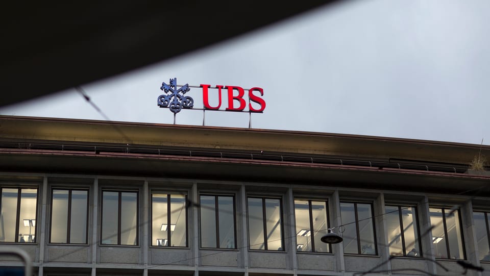 Il logo da la UBS sin il tetg da la sedia centrala da la banca a Turitg. Ins vesa las cazzolas da l'etascha il pli sisum dal bajetg èn illuminadas. Il tschiel è grisch. Il rest dal bajetg na vesan ins betg.