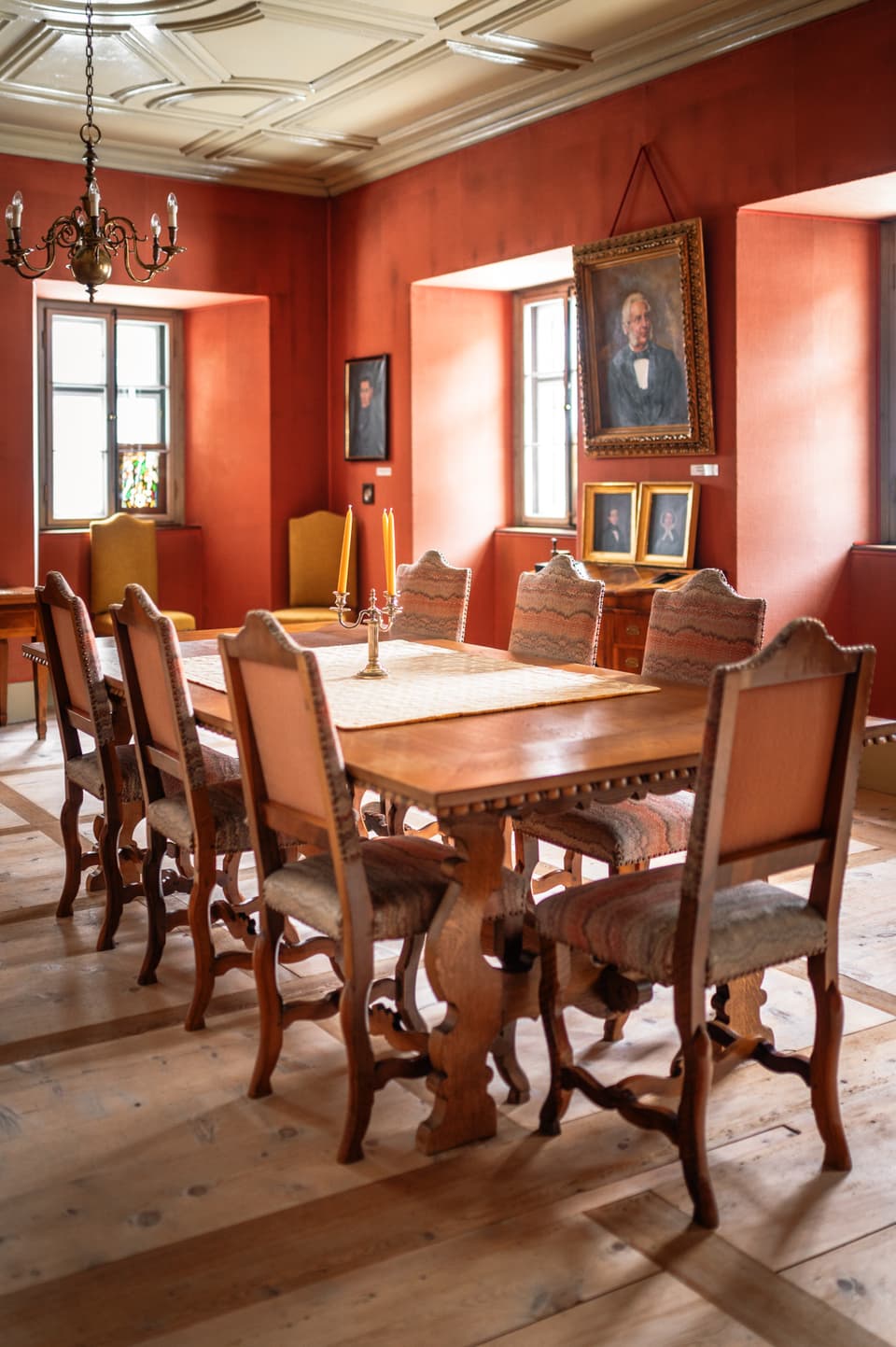 Im Zimmer mit oranger Tapete steht ein antiker Tisch und darum herum 7 mit Stoff bezogene Stühle.