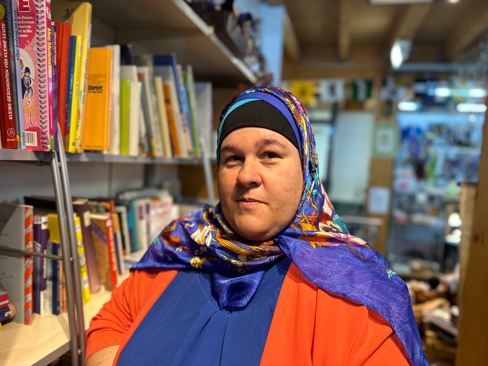 Frau mit blauem Kopftuch vor einem Büchergestell. 