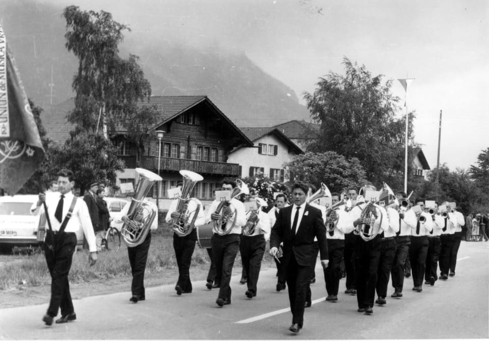 Fotografie s/w: Musikgesellschaft mit weissem Hemd und schwarzer Hose marschiert musizierend.