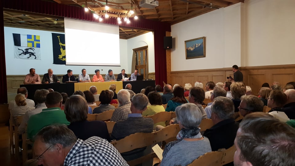 La suprastanza, 8 persunas sesan davos in lung pult sin il palc e guardan vers la glieud che sesa sin soptgas en la sala.
