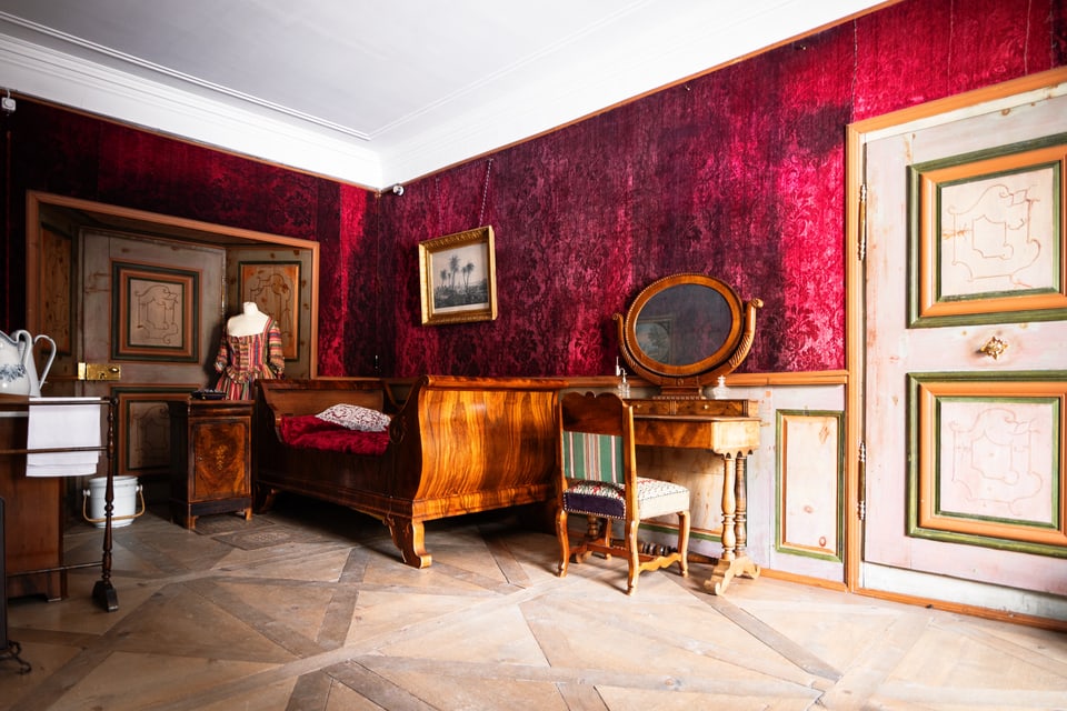 Ein Zimmer mit dunkelroter Tapete, einem alten Bett, Schminktisch mit Stuhl und einer verzierten Türe.