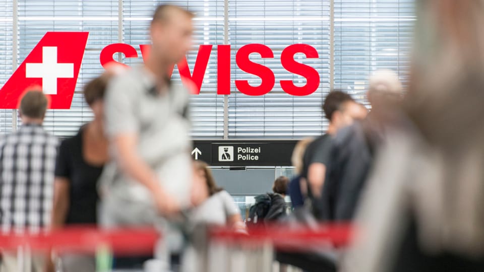 Passagiers a l'eroport a Turitg, en il fund il logo cotschen da la Swiss.