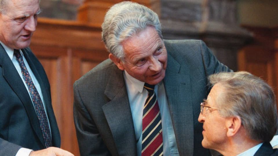 Paul Gemperli en discussiun cun dus umens cun vestgì e cravatta. 