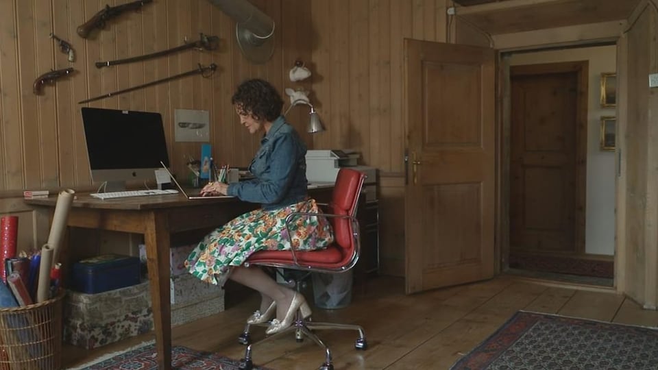 Romana Ganzoni en ses biro. Dunna en ina stiva. Sesa vid ina maisa davant il computer.