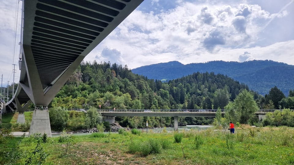 Im Vordergrund sind zwei Brücken, eine Weise und eine Frau zu sehen. Im Hintergrund ist eine Berglandschaft.