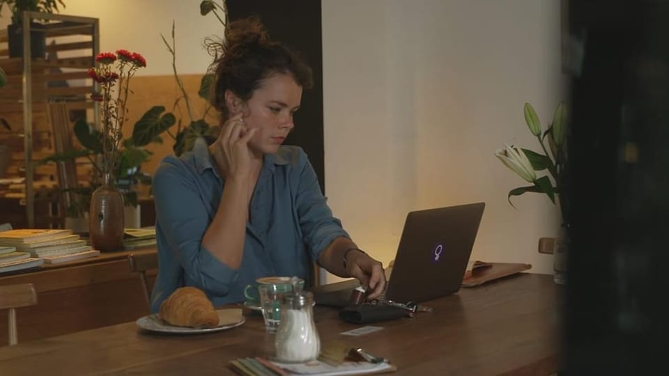 Viola Cadruvi cun computer, café e cornet. Dunna giuvna sesa vid ina maisa cun laptop e café davant ella.