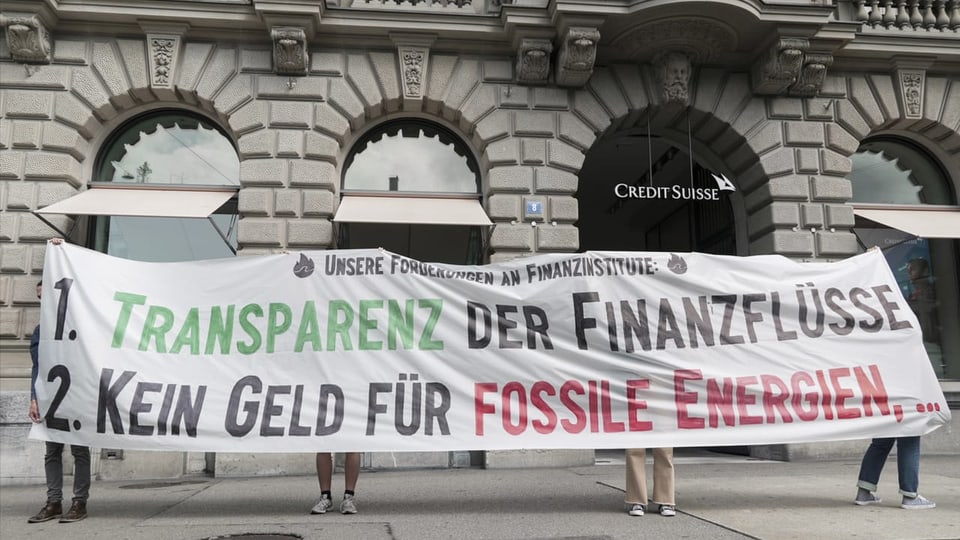 Quatter persunas tegnan in transparent cun scrit: «1. Transparenz der Finanzflüsse e 2. Kein Geld für Fossile Energien.