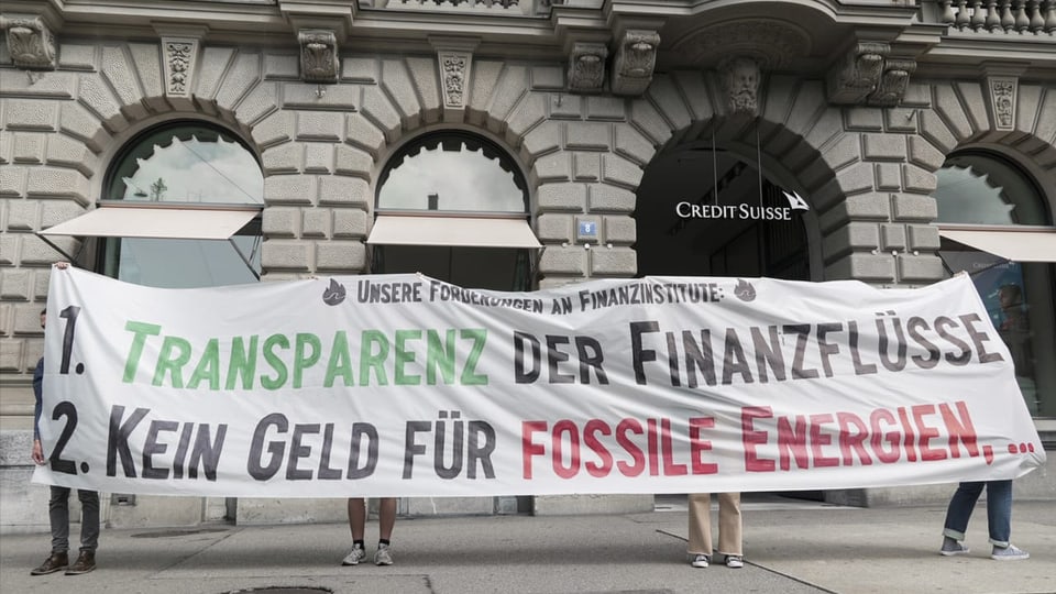 Quatter persunas tegnan in transparent cun scrit: «1. Transparenz der Finanzflüsse e 2. Kein Geld für Fossile Energien. 