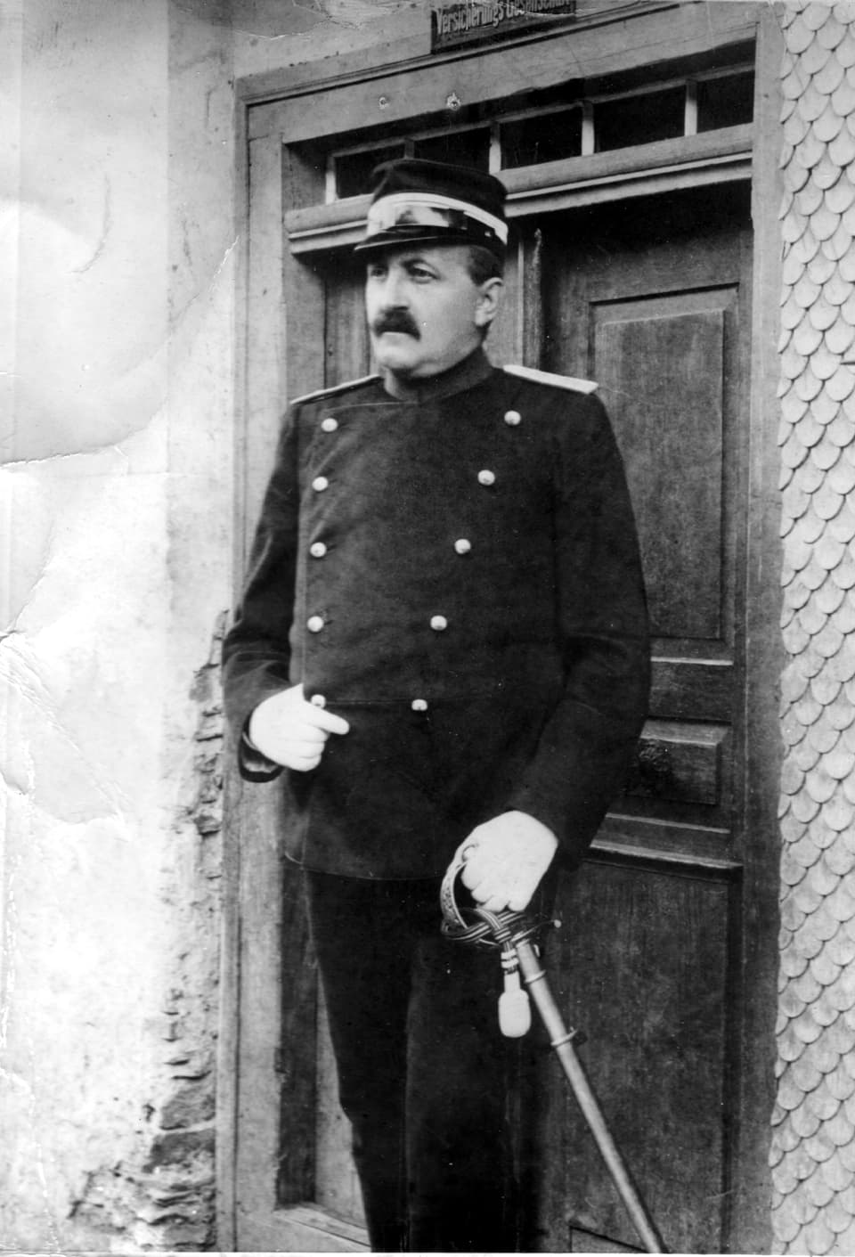 Fotografie s/w: Ein Mann mit Schnauz steht in Militäruniform und mit einem Degen vor einer Haustüre. 